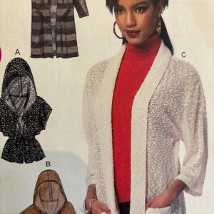 Peut inclure: Un cardigan blanc ouvert sur le devant avec des manches longues et des poches. Le cardigan est fait d'un tissu en tricot léger. Le mannequin porte un chemisier rouge à col roulé et un pantalon noir.
