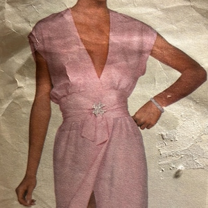 May include: A light pink dress with a plunging V-neckline and a wrap-style skirt. The dress features a jeweled belt buckle and a matching fabric belt. The dress has short sleeves and a high slit in the skirt.