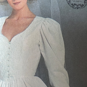 Puede incluir: Un vestido de novia blanco con escote de corazón y mangas largas. El vestido tiene un corpiño ajustado y una falda completa. El vestido está hecho de una tela suave y fluida. El vestido está adornado con un delicado encaje. El vestido se combina con un velo blanco. La imagen incluye el texto "Jessica, San Francisco".