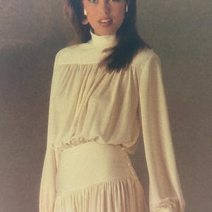 May include: Cream-colored dress with a high neckline and long sleeves. The dress features a gathered bodice, a fitted waist with button details, and a flowing skirt. The sleeves have a slight balloon shape at the wrist.