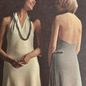 Può includere: Due donne in abiti vintage. Una indossa un abito all'americana color crema con una collana di perline scure. L'altra indossa un abito grigio senza schienale con un fiocco in vita. Lo sfondo è una superficie scura e strutturata.