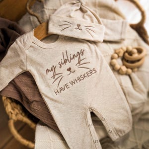 May include: A beige long-sleeve baby bodysuit and matching beanie set. The bodysuit features brown embroidery that reads "my siblings HAVE WHISKERS" with a cat nose and whisker design. The beanie also features an embroidered cat face.