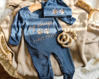 May include: A blue baby onesie and matching cap set with the text "my siblings HAVE PAWS" and paw print graphics. A wooden teething ring is also included. The set is displayed on a woven basket.