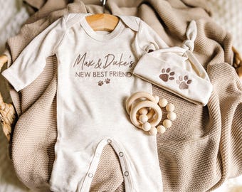 May include: A beige baby onesie, hat, and wooden rattle set. The onesie has the text "Max & Duke's NEW BEST FRIEND" with paw prints. The hat features embroidered paw prints. The rattle is made of wooden beads.