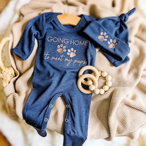 May include: A navy blue baby outfit set with the text "GOING HOME to meet my pups" and paw prints. The set includes a long-sleeved onesie, a knotted cap, and a headband, all with matching embroidered details. A wooden teether and a beaded rattle are also included.