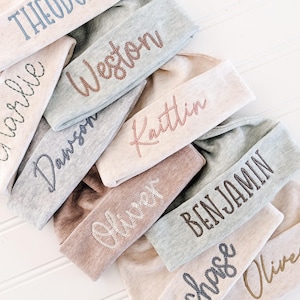 May include: A collection of personalized headbands in various neutral colors, each embroidered with a different name in a complementary thread color. The headbands are made of soft fabric and are arranged in a close-up shot.