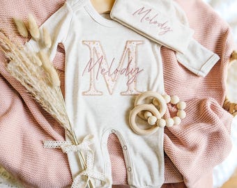May include: A baby gift set featuring a light gray onesie and matching cap, both embroidered with the name "Melody" in pink script. The onesie has a large letter "M" with a floral pattern. Includes wooden teething rings and a bouquet.