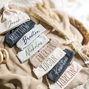 May include: A collection of personalized baby hats and headbands in various neutral colors. Each item is embroidered with a different name in a cursive font. The items are arranged on a textured, beige surface.