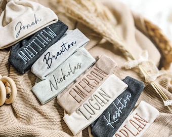 May include: A collection of infant knotted hats in various neutral colors including beige, charcoal gray, and light gray. Each hat features custom embroidery with names such as Jonah, Matthew, Braxton, Nicholas, Emerson, Logan, Kashton, and Harlan.