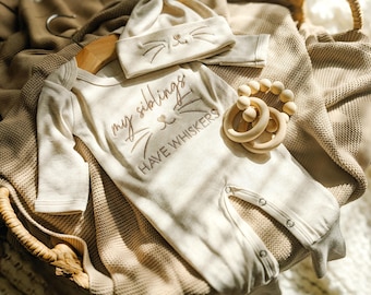 May include: A beige baby outfit set, including a long-sleeved onesie, a beanie, and a wooden teether. The onesie has the embroidered text "my siblings have whiskers" with a cat face design. The beanie also features an embroidered cat face.