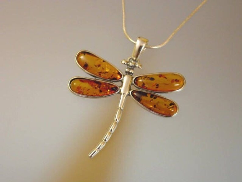 May include: A silver dragonfly pendant necklace with amber-colored wings. The dragonfly design features four amber-colored oval-shaped wings and a segmented body. The pendant hangs from a thin silver chain.