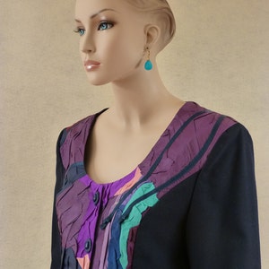 May include: A black blazer with a colourful, abstract patterned panel on the front. The panel features shades of purple, pink, green, and grey. The blazer has a button closure and long sleeves.