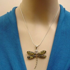 May include: A silver dragonfly pendant necklace. The pendant features a dragonfly design with four wings and a slender body. The wings are filled with a yellow-green material. The necklace is worn on a person wearing a blue top.