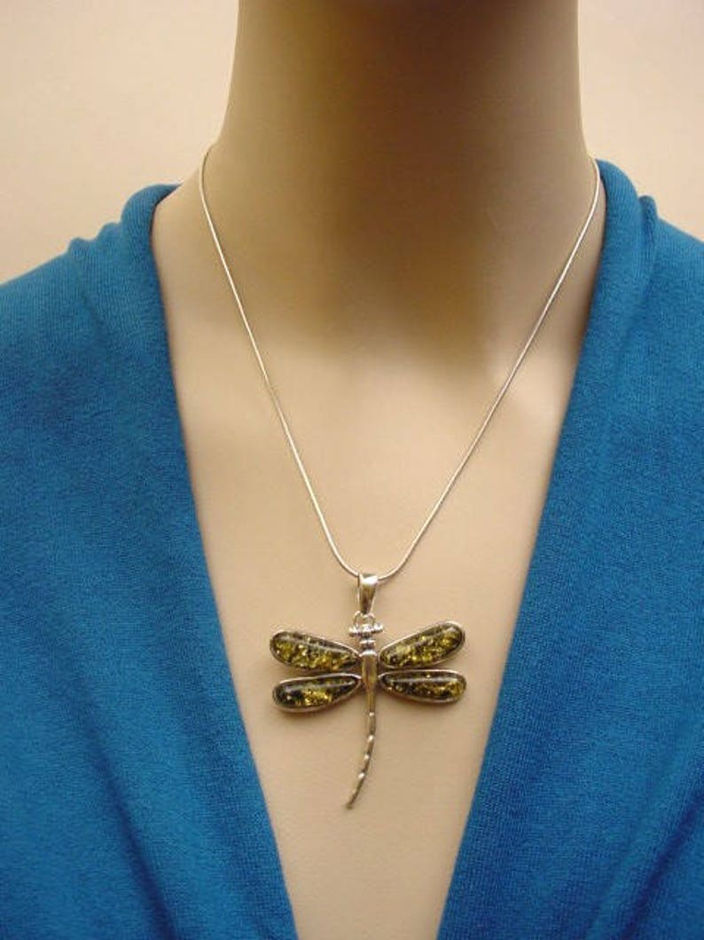 May include: A silver dragonfly pendant necklace. The pendant features a dragonfly design with four wings and a slender body. The wings are filled with a yellow-green material. The necklace is worn on a person wearing a blue top.
