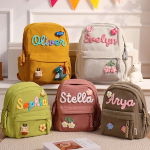 Peut inclure: Cinq sacs à dos en velours côtelé de différentes couleurs : jaune moutarde, gris clair, vert, rose et marron. Chaque sac à dos est personnalisé avec un nom en écriture cursive. Des appliqués décoratifs incluent des fleurs, un dinosaure et un papillon.