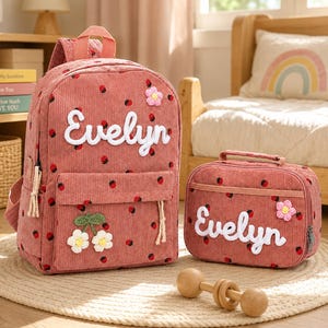 Könnte beinhalten: Ein Rucksack und eine passende Lunchtasche aus altrosa Kordstoff mit Erdbeermuster, dem Namen Evelyn in weißer Schreibschrift und kleinen gehäkelten Blumenapplikationen. Der Rucksack hat einen Tragegriff und eine Vordertasche, die Lunchtasche einen Henkel.