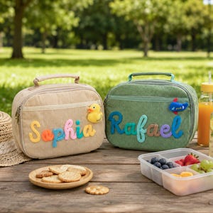 Pode incluir: Duas lancheiras retangulares de veludo cotelê com alças e zíperes. A bolsa bege apresenta o nome Sophia em letras de feltro coloridas e um patch de pato amarelo. A bolsa verde apresenta o nome Rafael e um patch de avião azul.