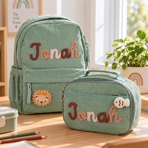 Puede incluir: Una mochila y una bolsa de almuerzo a juego en pana de color verde salvia, ambas personalizadas con el nombre Jonah bordado. La mochila incluye un parche de león y la bolsa de almuerzo tiene un pequeño adorno en forma de pez en la cremallera.