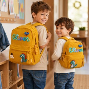 Peut inclure: Deux sacs à dos en velours côtelé jaune moutarde avec le prénom Oliver écrit en lettres cursives bleu sarcelle, un petit écusson d'avion bleu et un écusson de dinosaure vert. Chaque sac possède une poche frontale zippée et des bretelles.