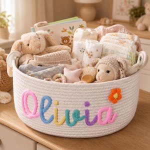 May include: A round white woven rope basket featuring the name Olivia embroidered in colourful yarn letters on the front, accented by a small orange flower. The basket is filled with baby items including plush toys, folded blankets, and nappies.