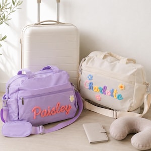 Peut inclure: Un sac de voyage violet clair avec le nom Paisley en orange, un sac de voyage crème avec le nom Charlotte en bleu, une valise à roulettes crème, un porte-passeport crème et un coussin de nuque crème, tous avec des petites fleurs.