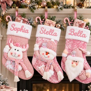 May include: Three pink Christmas stockings with white cuffs, each personalised with embroidered names Sophia, Stella, and Bella. The stockings feature plush reindeer, snowman, and Santa Claus appliqués, accented with candy canes and gift boxes.
