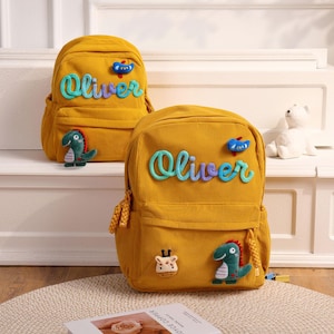 Op de afbeelding: Twee mosterdgele rugzakken van corduroy met de naam Oliver in blauwe en paarse schrijfletters. Elke tas is versierd met kleine pluche applicaties, waaronder een groene dinosaurus, een blauwe vogel en een giraffekop op het voorvak.