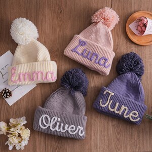 May include: Three knitted hats with pom-poms, each with a different name embroidered on the front: Emma, Luna, and June. The hats are in shades of pink, purple, and gray.