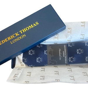 May include: A navy blue gift box with gold lettering reading "FREDERICK THOMAS LONDON". Inside, a navy blue tie with a white pattern is wrapped in tissue paper. The tissue paper is printed with the letters "FT".