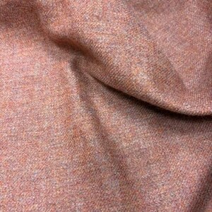 May include: A textured fabric in a warm, earthy terracotta orange hue. The material features a dense, woven appearance with subtle flecks of lighter and darker tones throughout, creating a complex, multi-dimensional surface with visible folds.