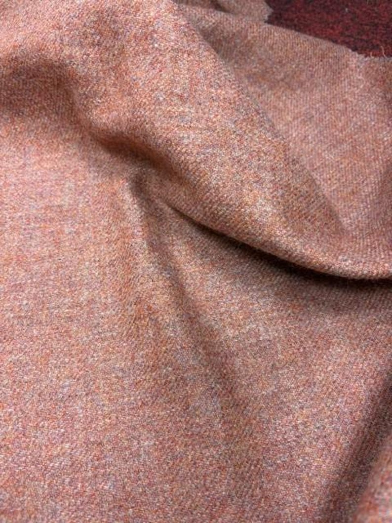 May include: A textured fabric in a warm, earthy terracotta orange hue. The material features a dense, woven appearance with subtle flecks of lighter and darker tones throughout, creating a complex, multi-dimensional surface with visible folds.