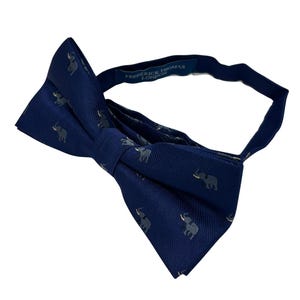 May include: A navy blue bow tie with a repeating pattern of light grey elephants. The bow tie has an adjustable strap with the text "FREDERICK THOMAS LONDON" printed on it.