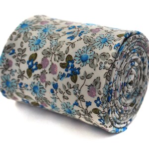 Puede incluir: Corbata enrollada con un estampado floral. La corbata presenta un fondo blanco con un patrón repetido de flores y hojas azules, celeste, verdes y moradas. La corbata está enrollada, mostrando el patrón en los bordes.