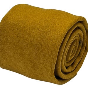 May include: Rolled mustard-colored necktie. The tie is made of a textured fabric and is tightly rolled, showcasing its solid color and material. The end of the tie is visible, revealing the fabric's weave.