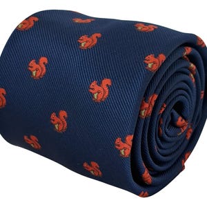 Può includere: Una cravatta blu navy con un motivo ripetuto di piccoli scoiattoli rosso-arancio, ognuno dei quali tiene in mano una minuscola noce verde. Il tessuto presenta una visibile trama diagonale a saia ed è arrotolato in forma cilindrica.
