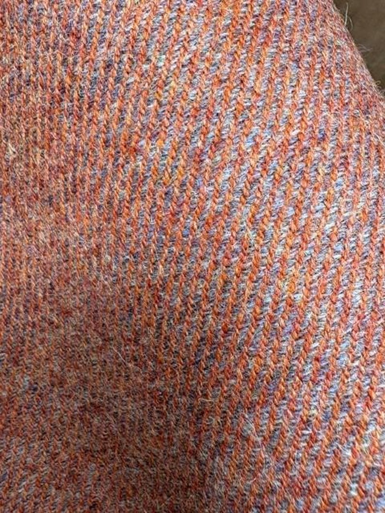 May include: A close-up view of a woven textile featuring a diagonal twill pattern. The fabric is composed of alternating threads in vibrant orange and soft lavender, creating a textured, multi-colored appearance with a visible fiber structure.
