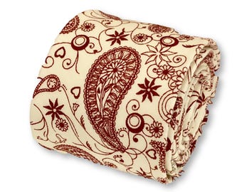 Pode incluir: Cobertor creme dobrado com um padrão paisley bordô. O design inclui grandes formas paisley, flores e vinhas em espiral. O cobertor está cuidadosamente dobrado, mostrando os intrincados detalhes do tecido.