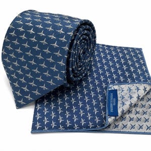 Puede incluir: Una corbata azul enrollada y un pañuelo de bolsillo cuadrado a juego, ambos con un patrón repetitivo de pequeños aviones de combate plateados. El pañuelo incluye una pequeña etiqueta de tela azul con el texto blanco "FIGHTER JET COLLECTION".