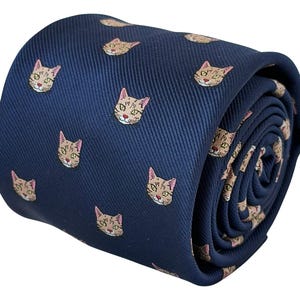 Può includere: Una cravatta blu navy con un motivo ripetuto di piccoli volti di gatto ricamati in beige e marrone. Il tessuto presenta una visibile trama diagonale a saia e la cravatta è arrotolata in forma cilindrica, mostrando la stampa dei gatti.