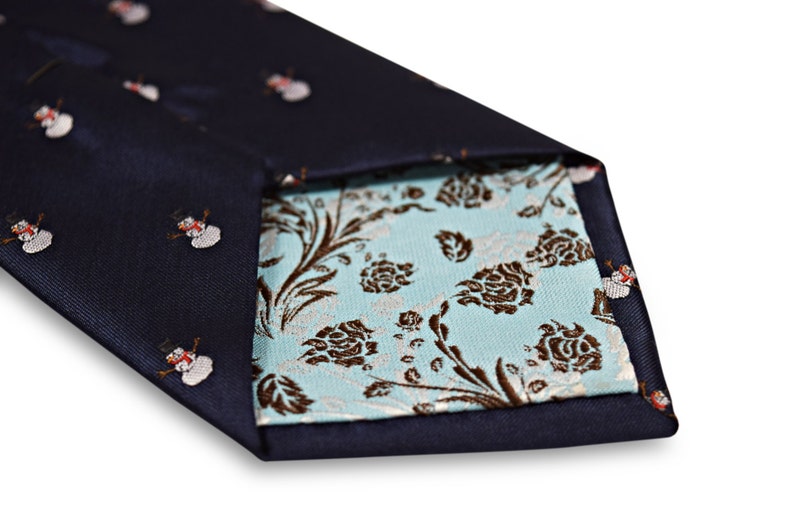Puede incluir: Una corbata azul marino con un patr&oacute;n de mu&ntilde;eco de nieve blanco. La corbata es reversible y tiene un patr&oacute;n floral azul claro en el otro lado.