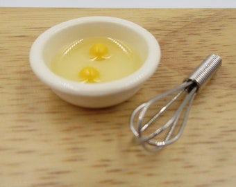 May include: A white bowl with two yellow egg yolks in a clear liquid, sitting on a wooden surface next to a silver whisk.