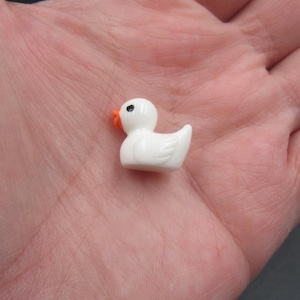 May include: A small white ceramic duck figurine with an orange beak and black eyes. The duck is sitting on a hand.