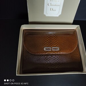 Peut inclure: Une pochette à la texture peau de serpent marron avec un logo CD argenté sur le rabat avant, présentée dans une boîte beige. Le couvercle de la boîte comporte une étiquette blanche avec le texte 'les sacs Christian Dior' en police serif.