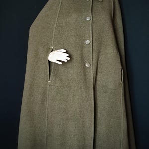 Pode incluir: Capa verde-oliva com gola redonda e fecho de botões. A capa apresenta dois bolsos frontais e uma silhueta fluida. A peça de roupa é exibida num manequim sobre um fundo escuro.