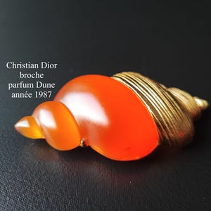 May include: An orange and gold seashell brooch. The brooch is in the shape of a spiral seashell with a gold-coloured spiral end. The text on the image reads "Christian Dior broche parfum Dune année 1987."
