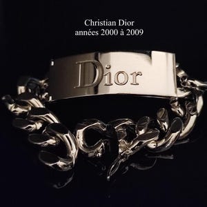 Op de afbeelding: Zilverkleurige armband met een Dior-plaat en een dikke schakelketting. De naam "Dior" is prominent aanwezig. De tekst "Christian Dior années 2000 à 2009" is zichtbaar.