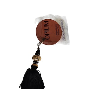 May include: A round, brown tag with the word "OPIUM" printed in white. The tag is attached to a black tassel with a gold and black bead. The tag also has the text "Parfum de Parfum" and "Perfumed Tassel" printed in white.