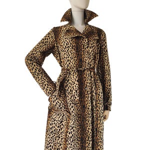 May include: A knee-length trench coat featuring a tan and black leopard print pattern. The coat includes a pointed collar, long sleeves, a double-breasted button front closure, and a matching fabric tie belt at the waist to secure the garment.