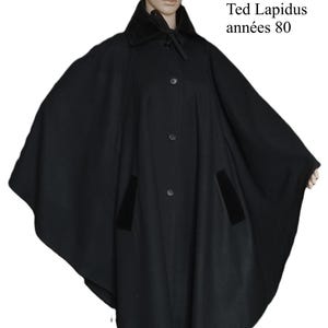 Op de afbeelding: Een zwarte cape met een puntige zoom, een hoge kraag en een knoopsluiting aan de voorzijde. Het kledingstuk heeft twee verticale zakken met donkere randen en bevat de tekst Ted Lapidus années 80 aan de zijkant. Het ontwerp is wijd en vloeiend.