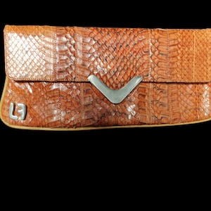 Op de afbeelding: Een bruine clutch van slangenleer met een zilveren sluiting. De clutch heeft een klein zilveren label met de letter "L" erop.